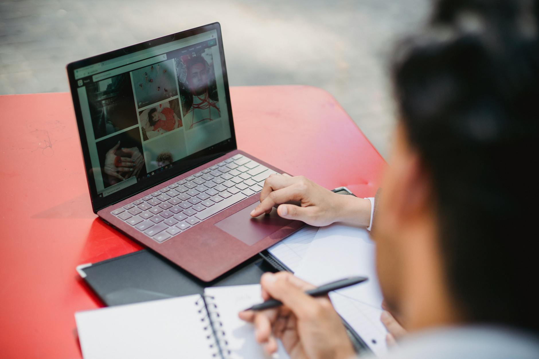 Individual working on a laptop and writing notes on a red table outdoors.