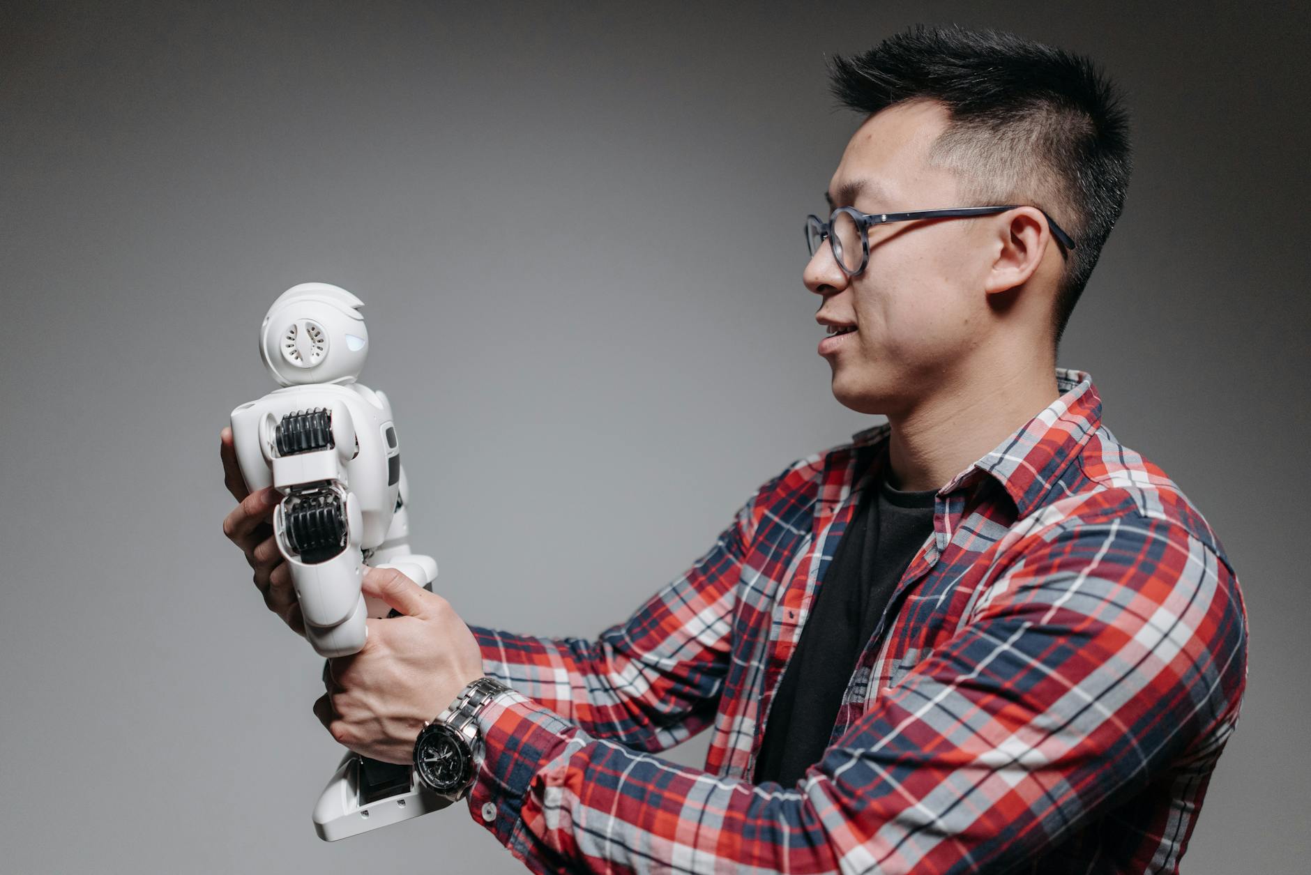 Asian man with glasses in plaid shirt holding a robot toy indoors. Studio shot.