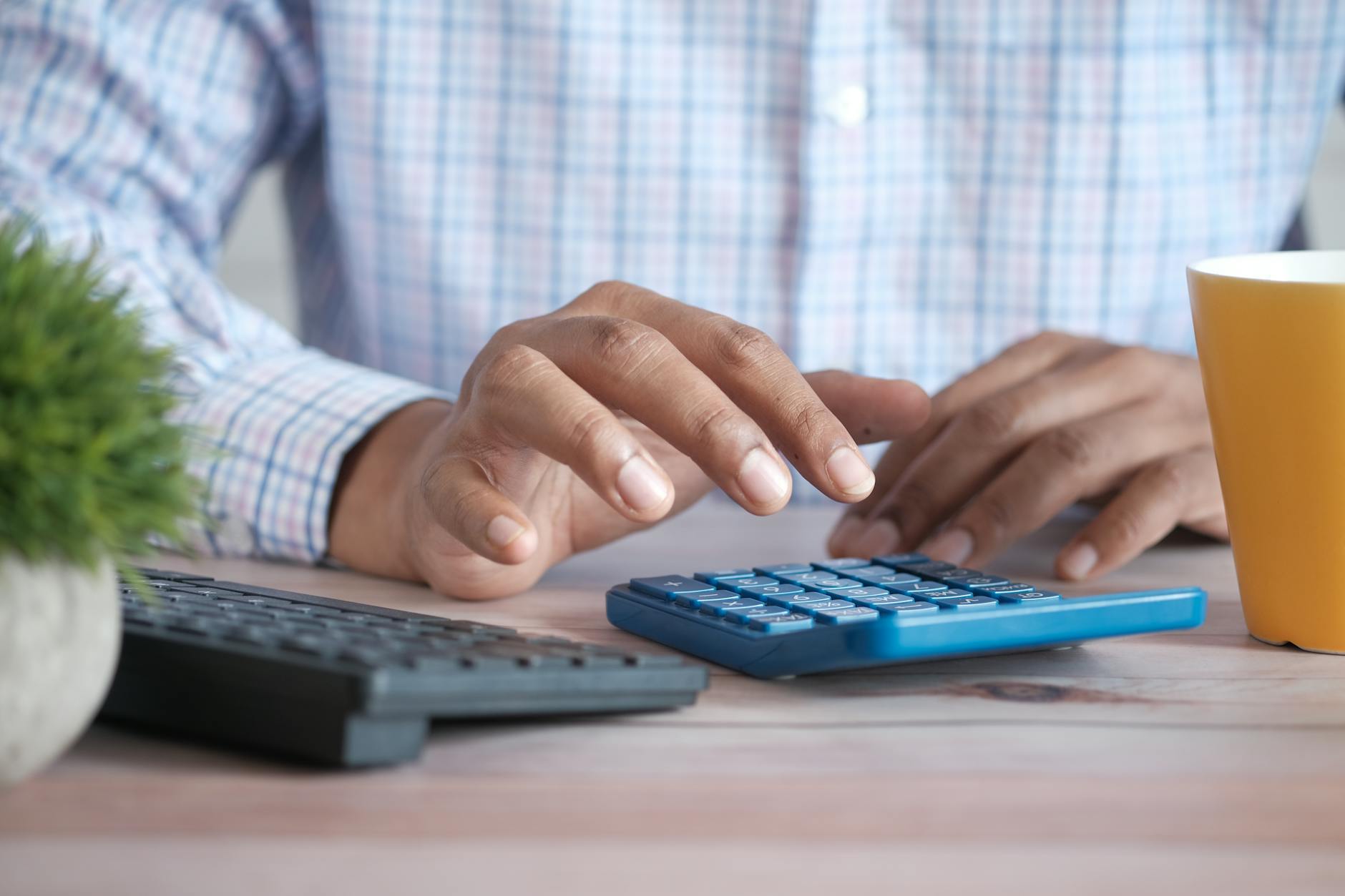A person using a blue calculator on a wooden desk with a keyboard and plant nearby.