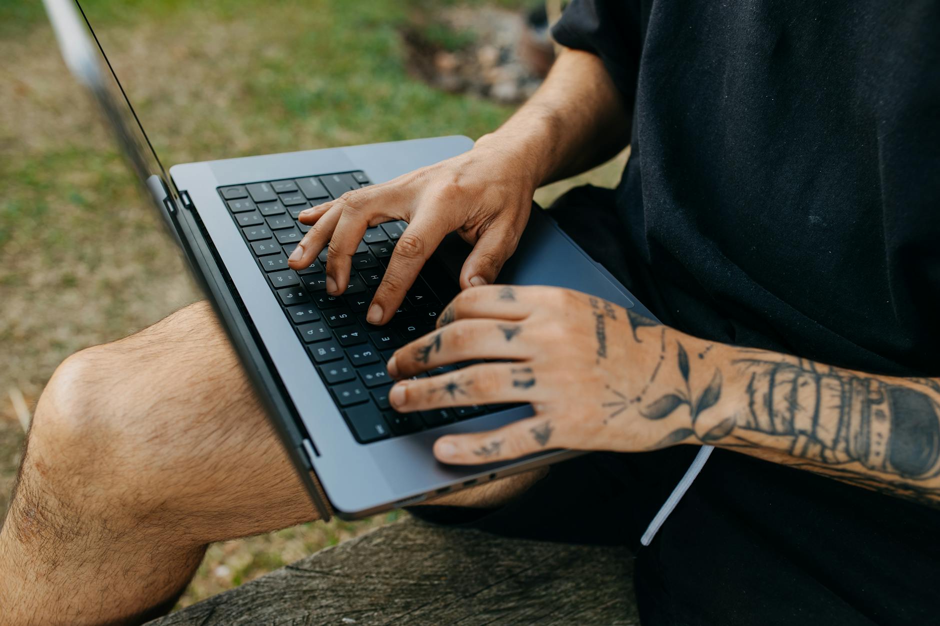 A tattooed man types on a laptop while sitting on a bench outside, surrounded by grass.