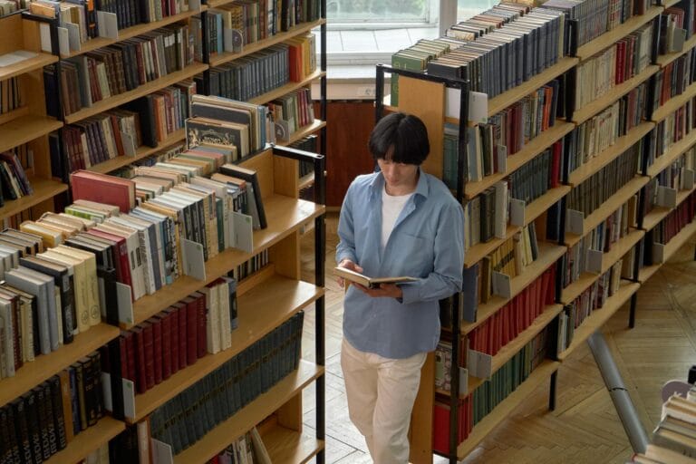 A young man reads intently in a bright library surrounded by bookshelves.