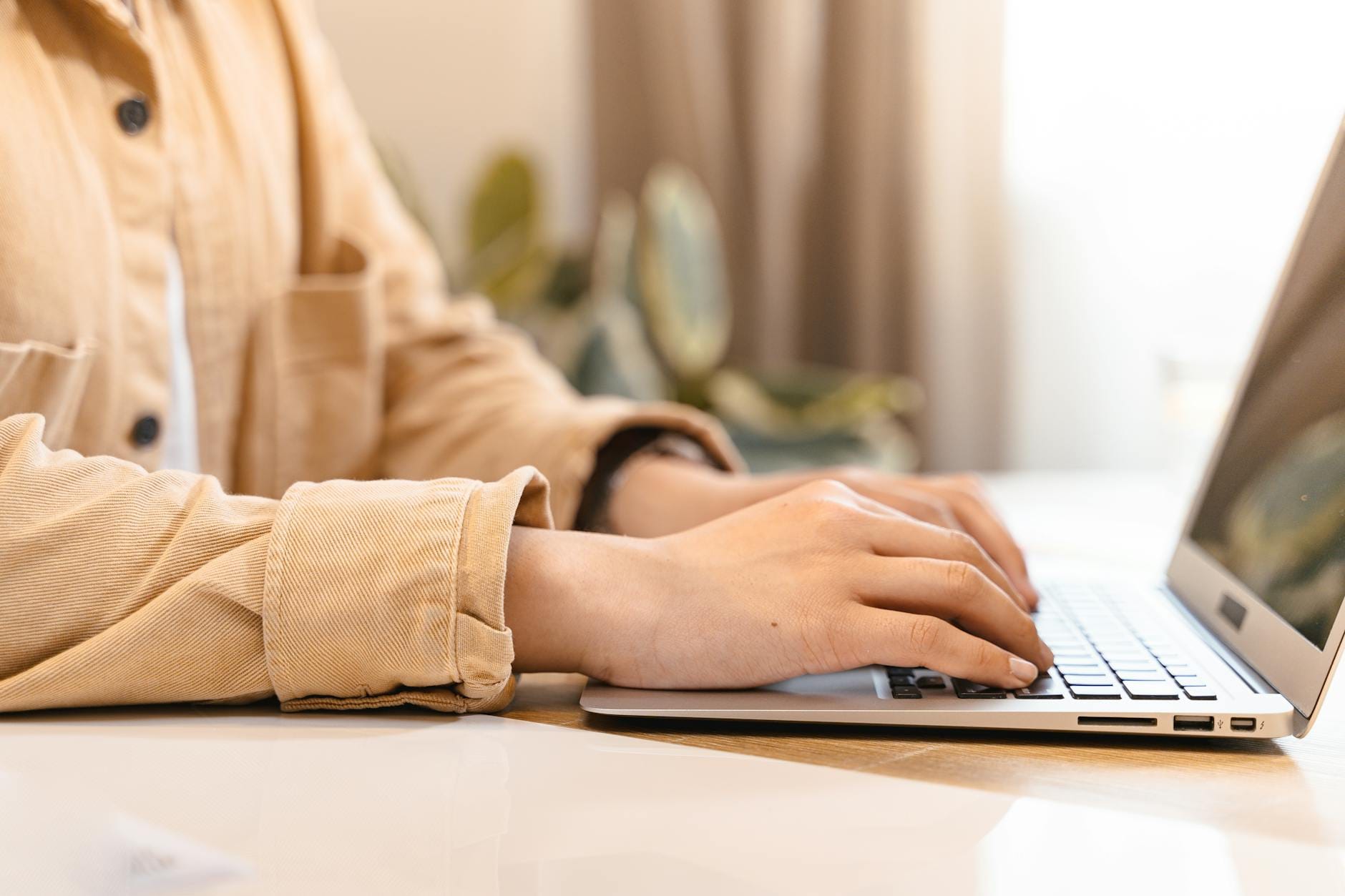 Close-up of hands typing on a laptop indoors, wearing a casual jacket.