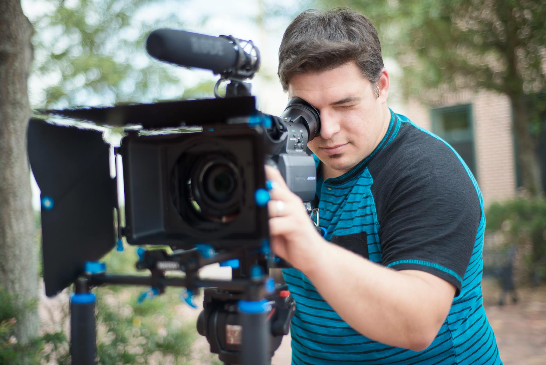 Young male videographer using a professional video camera outdoors with a blurred background.