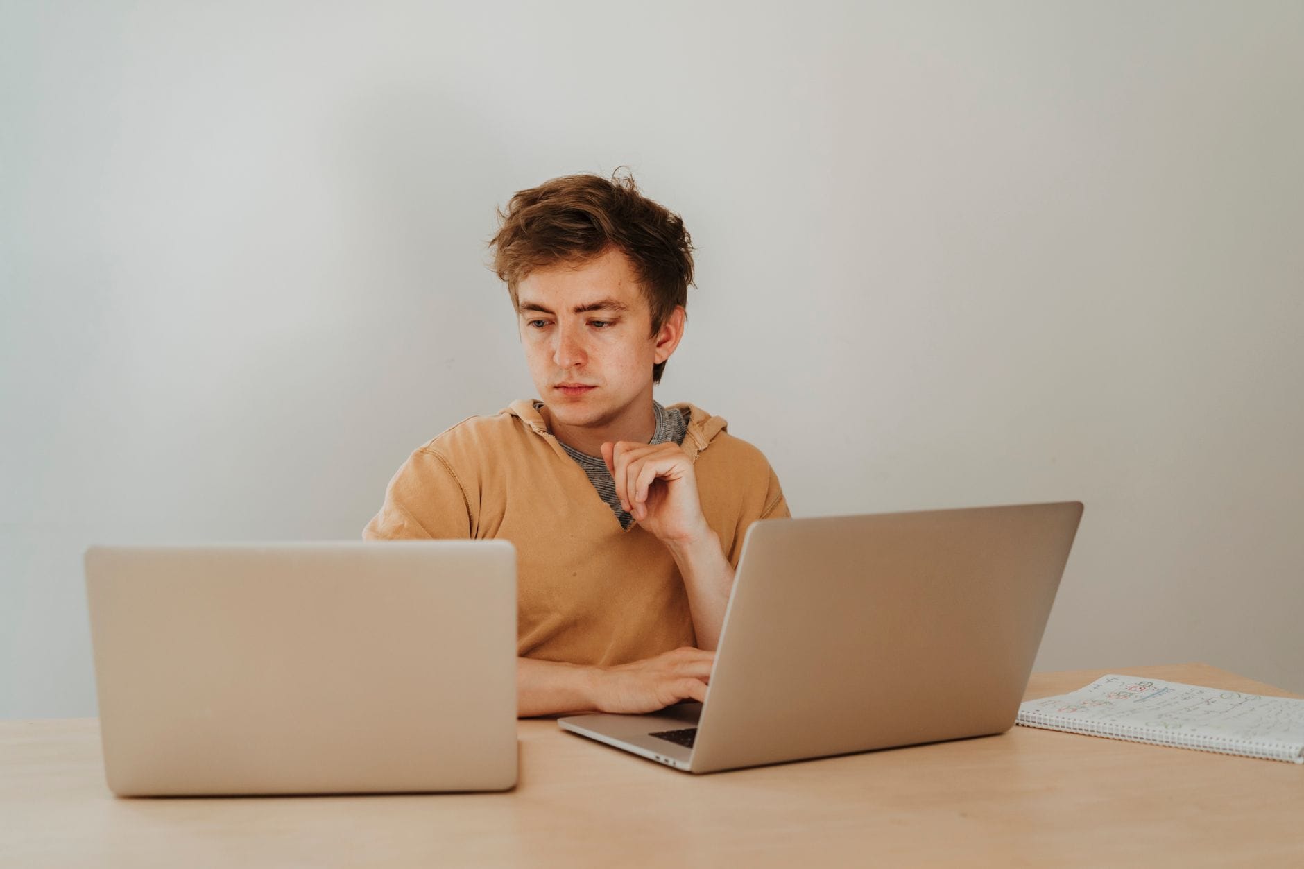 Focused young man working on two laptops in a home office setting, with a notebook visible on the desk. — cursor review 