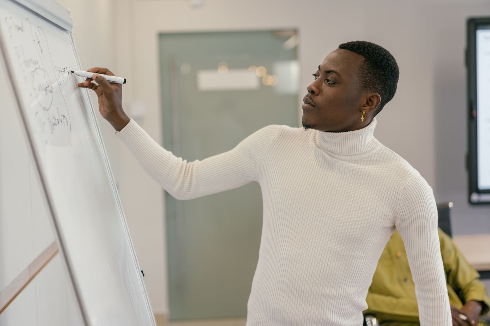 A young man in a white turtleneck writes on a whiteboard in a modern office setting. — gamma app review 2026
