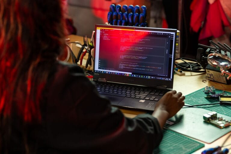A software developer writing code on a laptop at a cluttered workbench with electronic tools. — claude mania enterprise