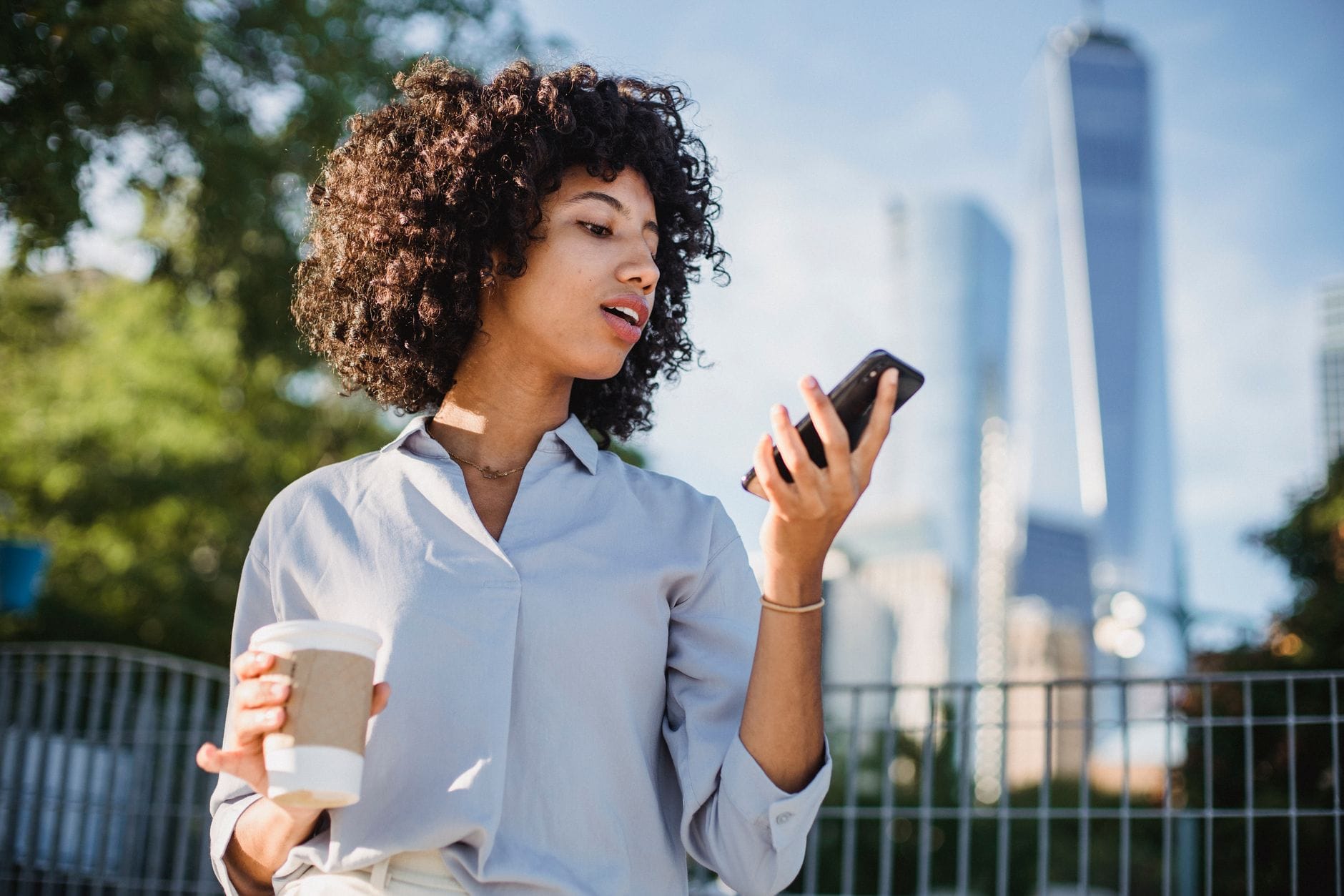 Young woman with curly hair uses smartphone outdoors, holding coffee. — new siri release date iphone 2026